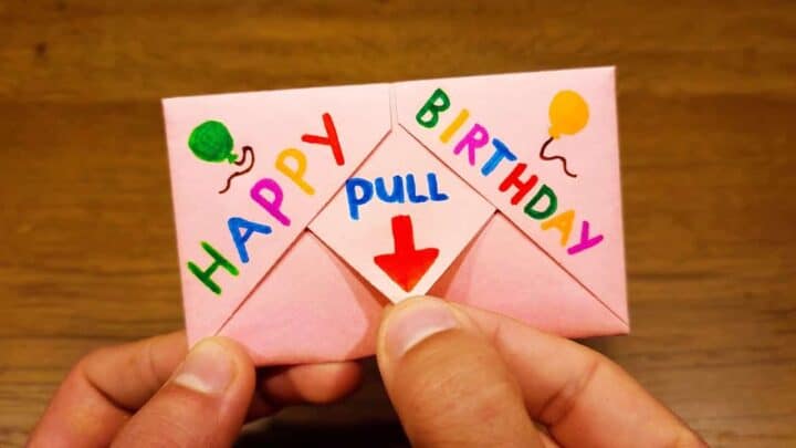 Hand holding a pink birthday card with a pull tab and colorful 'HAPPY BIRTHDAY' message.