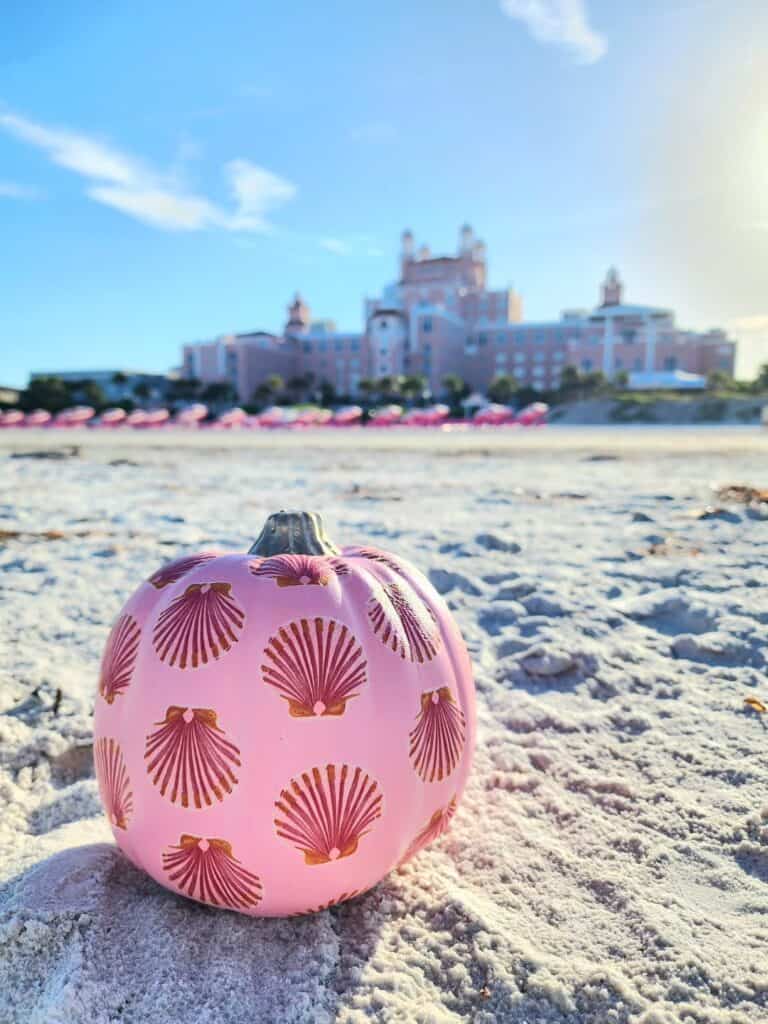 Pink pumpkin with shell pattern on sandy beach with a hotel in the background.